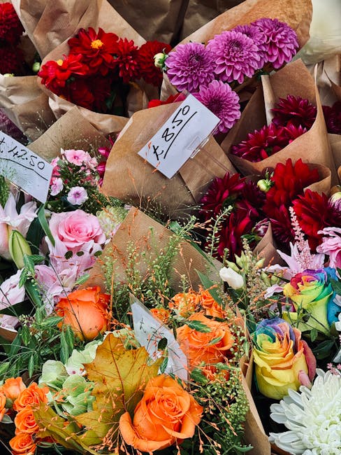A vibrant display of fresh floral arrangements featuring a variety of flowers including pink roses, orange roses, red dahlias, purple chrysanthemums, and multicolored roses, all wrapped in brown kraft