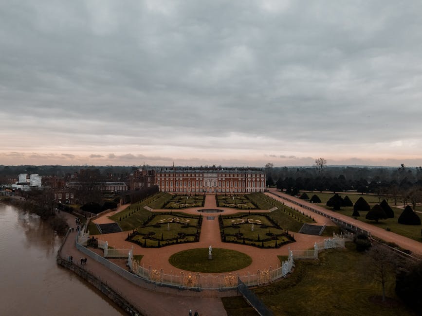 Aerial view of Syon Park featuring a grand historic mansion at the center, surrounded by meticulously landscaped formal gardens with geometric flower beds and neatly trimmed hedges. The gardens are ar