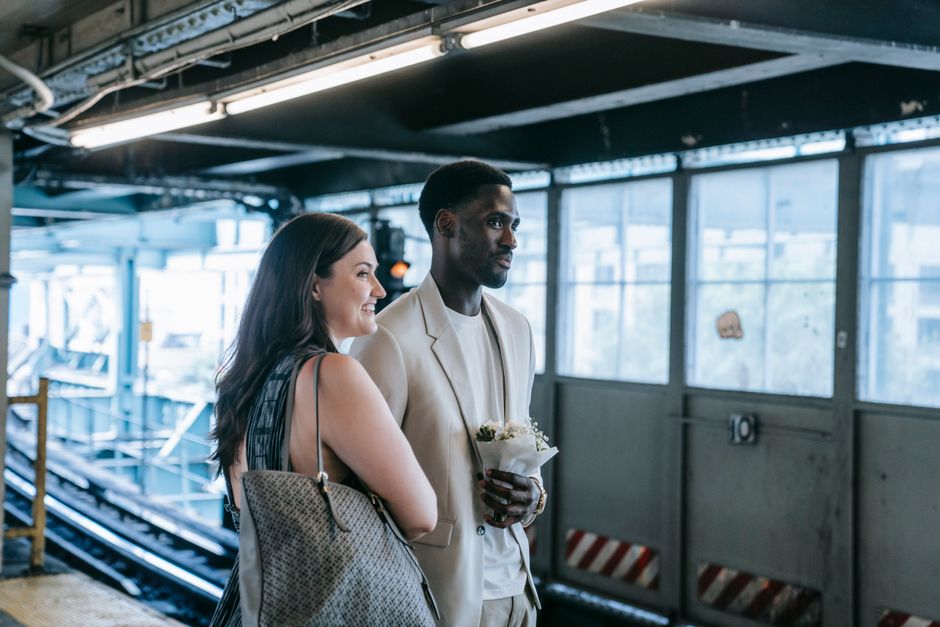 A woman and a man stand together on an indoor platform at Isleworth Station, near a set of railway tracks visible in the background. The woman, with long brown hair, is smiling and holding a patterned