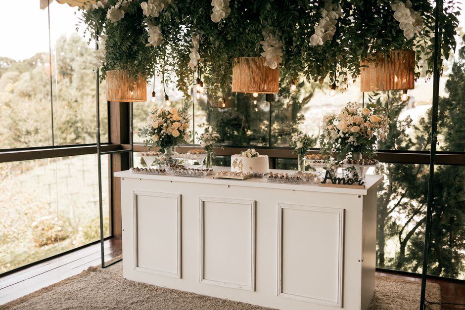 A white wooden sideboard decorated with multiple floral arrangements in glass vases, featuring soft pastel-colored roses, white hydrangeas, and greenery. Above the sideboard, three hanging woven ratta