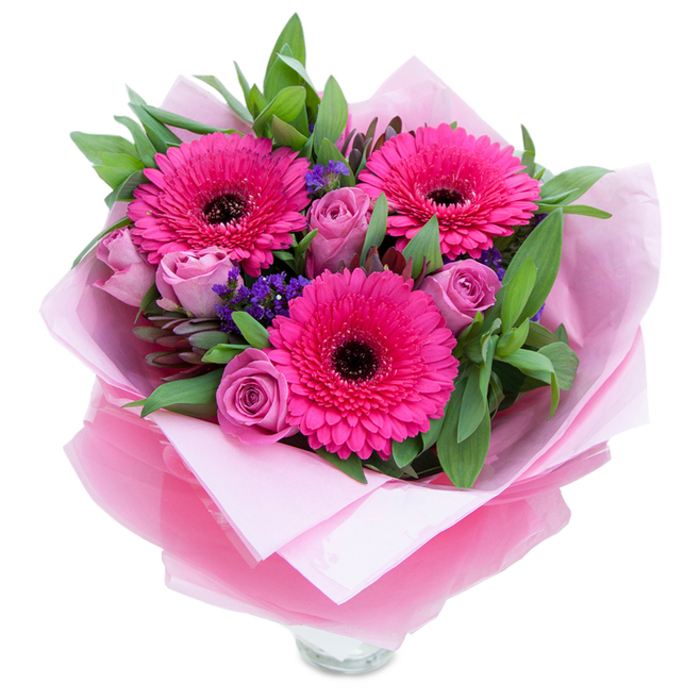 I see a bouquet that balances playfulness and refinement: three bold gerberas in a vivid fuchsia command attention with their wide faces and dark, contrasting centers, while clusters of soft lavender-pink roses weave a romantic softness through the arrangement. Between these principal blooms, tiny clusters of deep purple statice add a sprinkling of cool color and a textured, slightly papery contrast that lifts the eye. Lush green foliage-broad leaves that read as salal or eucalyptus-frames each bloom, their glossy surfaces and subtle veining giving the bouquet body and a garden-fresh aroma. The flowers are wrapped with a florist's tenderness: a gentle layer of matte pale pink tissue provides a muted, warm base, while an outer sheet of translucent fuchsia-pink cellophane, slightly crinkled, contributes a faint shimmer and ties the whole colour story together. The lower portion reveals the rim of a clear glass vase, hinting at stems trimmed for longevity and an arrangement meant for immediate display. Under soft, even lighting the petals show their textures-the velvety rose curves, the crisp gerbera rays, the tiny clustered statice-inviting touch and close inspection. The overall feeling is intimate and celebratory, perfect for marking a birthday, Mother's Day, or sending a thoughtful note to a friend across Isleworth High Street or to a neighbour returning from a walk in Syon Park. This bouquet reflects careful technique and a dedication to freshness that a local florist would be proud to deliver.